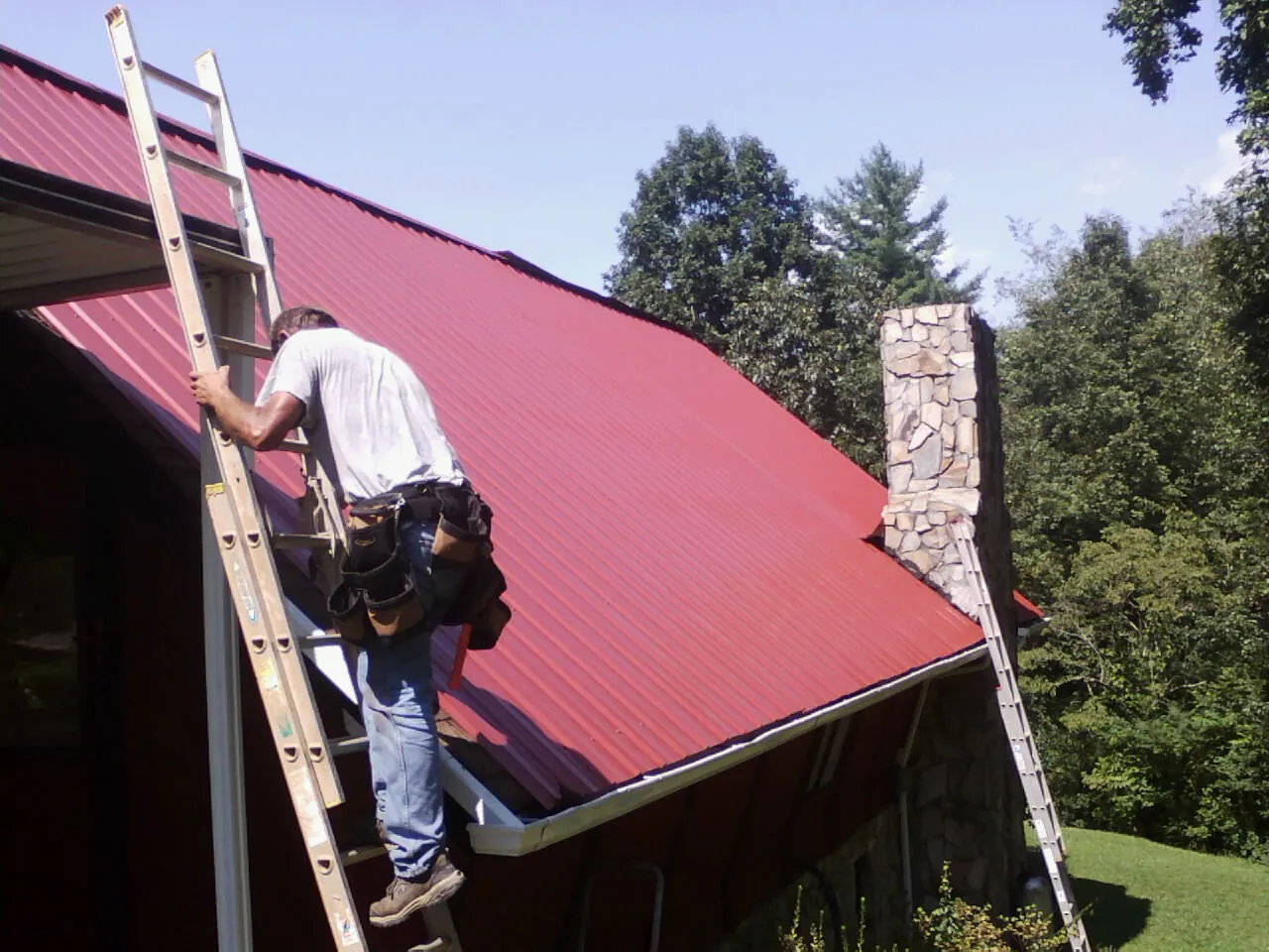 Roofer on a ladder beside a red metal roof and stone chimney — Soffit & Fascia Repair by Roofing Brothers in Tidewater
